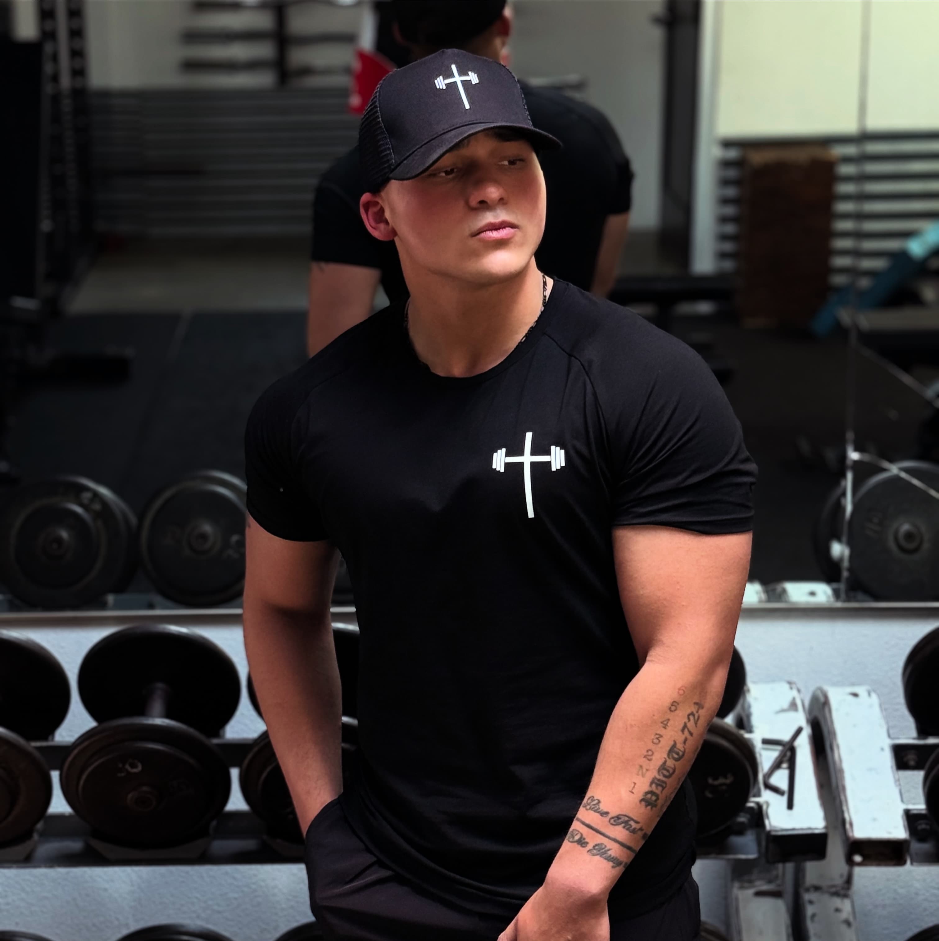 Christian Baughman in a black tee and cap by the dumbbell rack